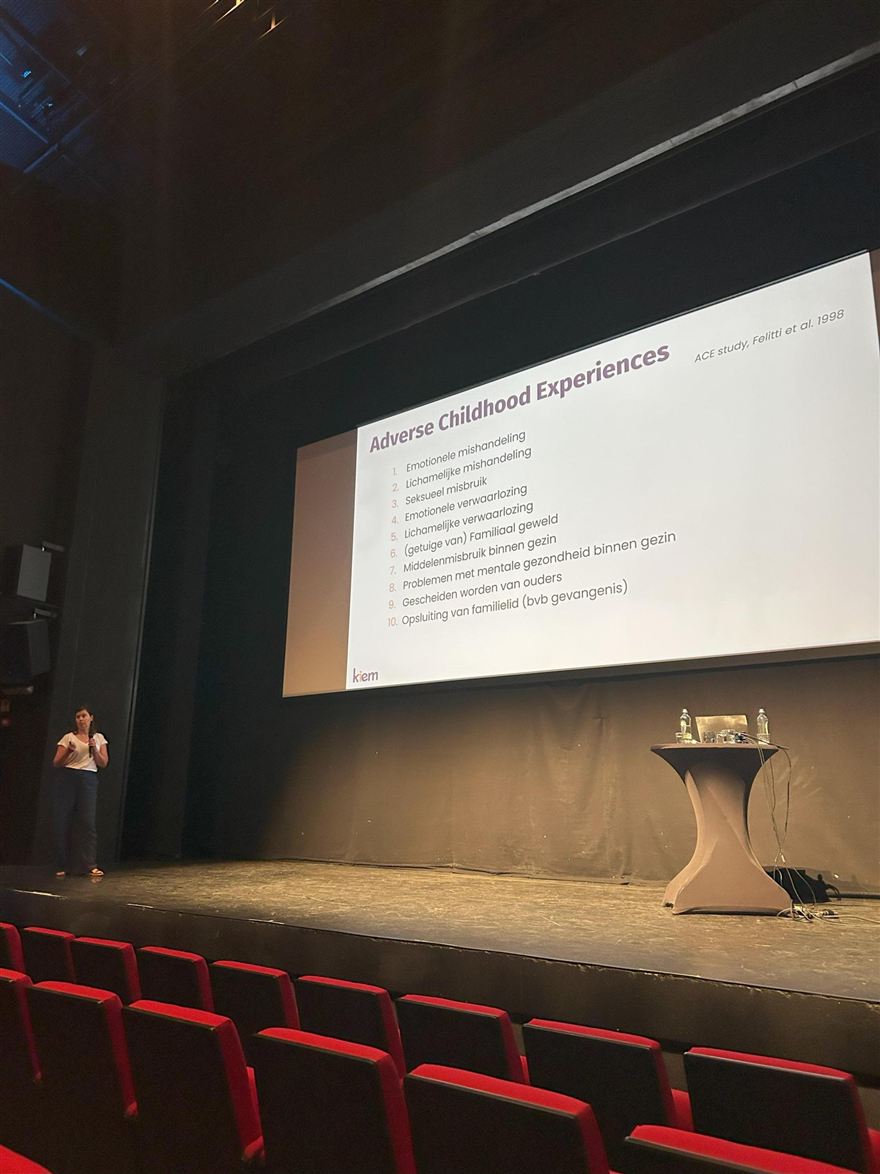 Spreekster op het podium naast een dia over Adverse Childhood Experiences.