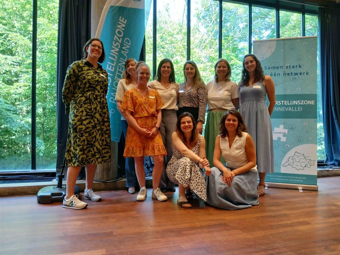 Groepsfoto van acht vrouwen tussen de banners van Eerstelijnszone Pajottenland en Eerstelijnszone Zennevallei.