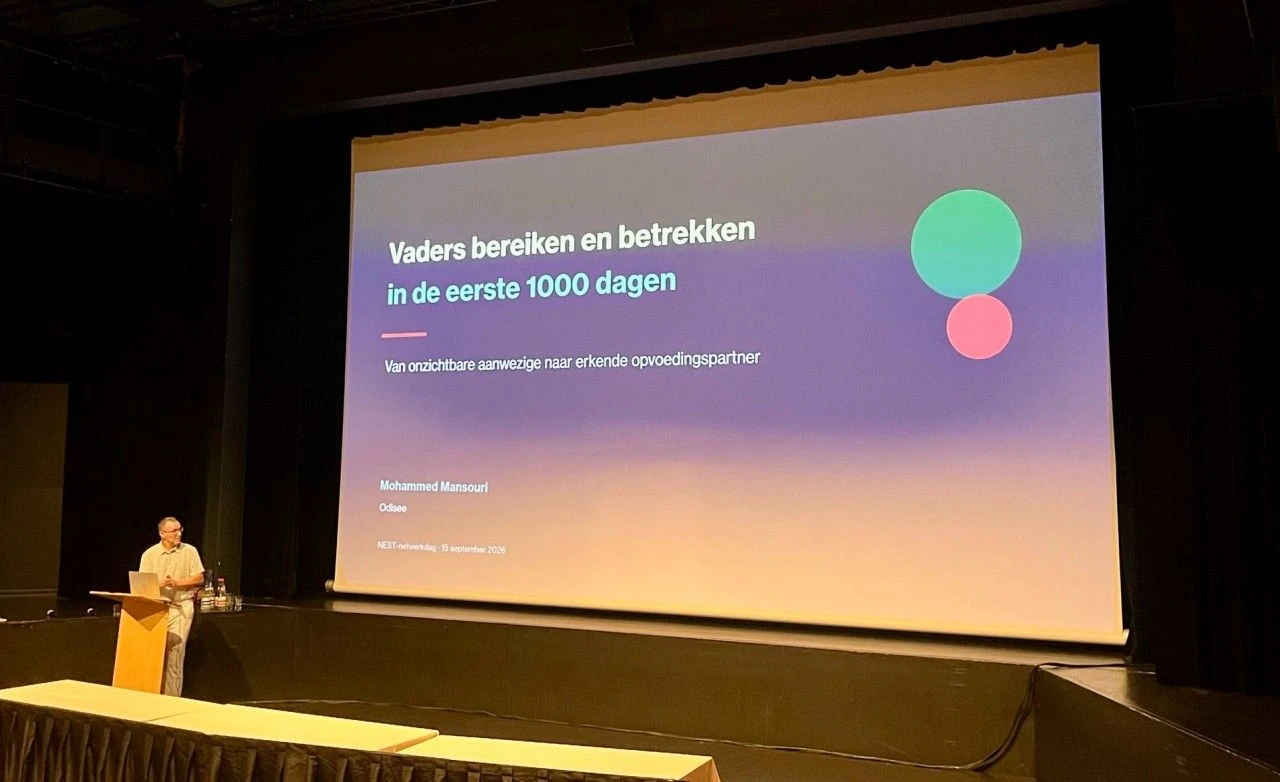 Spreker naast een groot scherm met de titel 'Vaders bereiken en betrekken in de eerste 1000 dagen'.