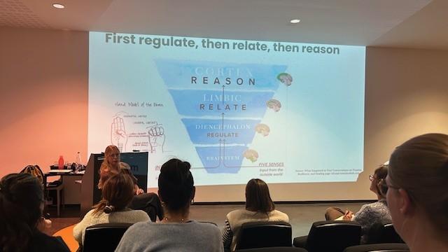 Het publiek kijkt naar een dia met de titel 'First regulate, then relate, then reason'.